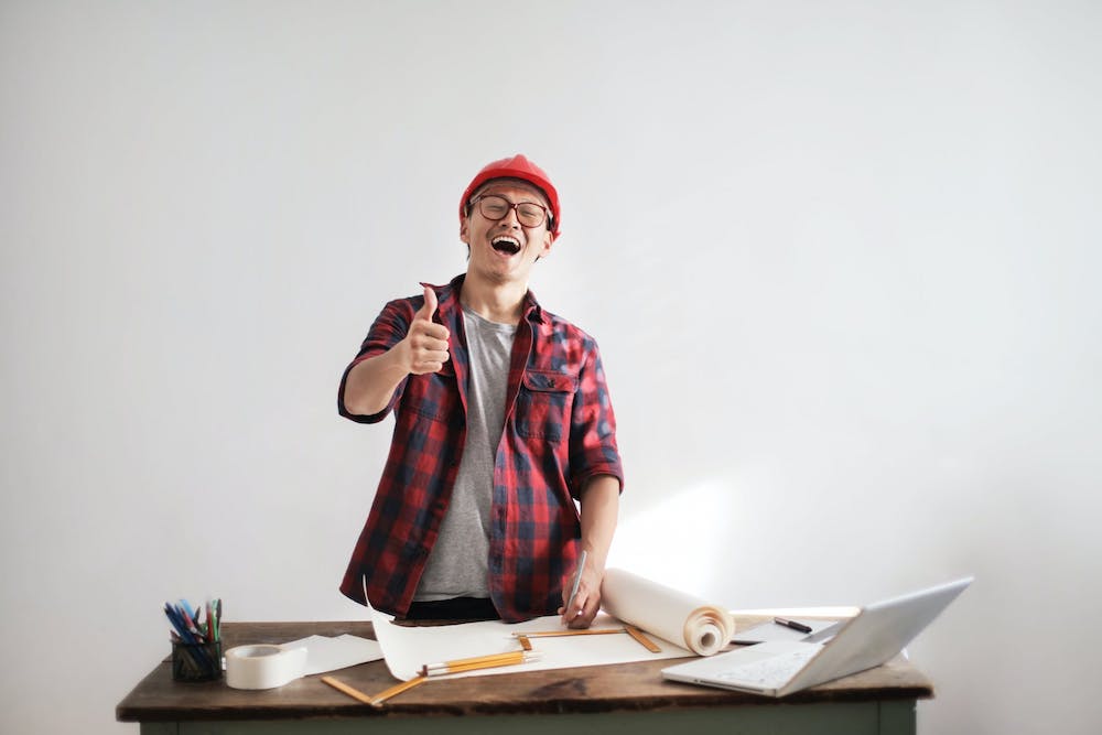Na zdjęciu uśmiechnięty, młody mężczyzna w koszuli w kratę i kasku na głowie. Trzyma kciuk podniesiony do góry. Przed sobą, na biurku, ma rozłożone kartki, ołówki oraz laptop.